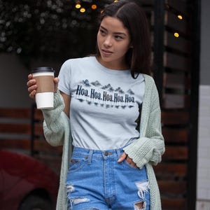 May include: A light blue short-sleeve t-shirt featuring a graphic print of dark clouds and trees above the repeated text "Hoa, Hoa, Hoa, Hoa, Hoa". The shirt is paired with blue denim shorts and a long, light green open-knit cardigan sweater.