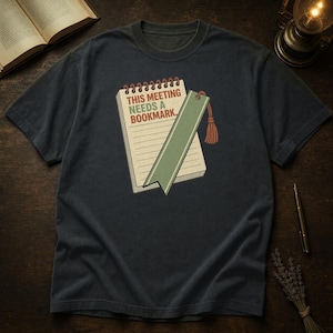 Puede incluir: Una camiseta de manga corta gris carbón oscuro con un gráfico de un bloc de notas de espiral, un marcapáginas verde y una borla. El bloc de notas muestra el texto "THIS MEETING NEEDS A BOOKMARK." en letras rojas y verdes sobre un fondo crema.