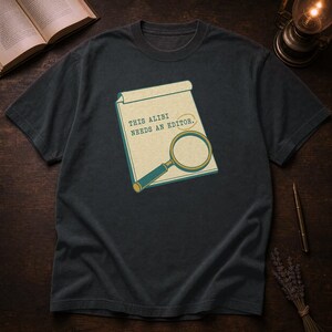 Puede incluir: Una camiseta gris carbón de manga corta con un gráfico de un bloc de notas y una lupa. El bloc de notas muestra el texto "THIS ALIBI NEEDS AN EDITOR." en una fuente estilo máquina de escribir, con un círculo alrededor de la palabra "EDITOR."