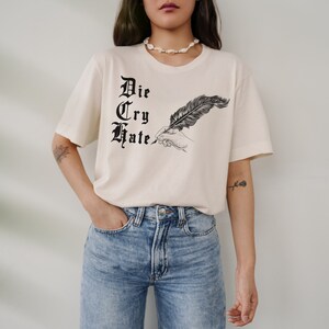May include: A cream colored t-shirt with a black graphic of a hand holding a feather pen and the words "Die Cry Hate" in a gothic font.