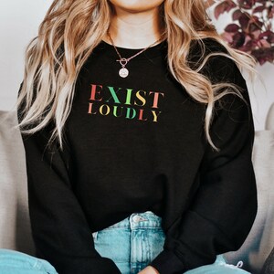 May include: A black sweatshirt with the text "EXIST LOUDLY" in rainbow colors. The sweatshirt is worn by a person with long blonde hair and blue jeans.