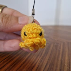 Könnte beinhalten: Ein kleiner, leuchtend gelber, gehäkelter Oktopus-Anhänger mit rundem Kopf und mehreren geringelten Tentakeln. Der Oktopus hat zwei kleine, schwarze gestickte Augen und ist an einer dünnen schwarzen Kordel mit silberfarbenem Metallverschluss befestigt.