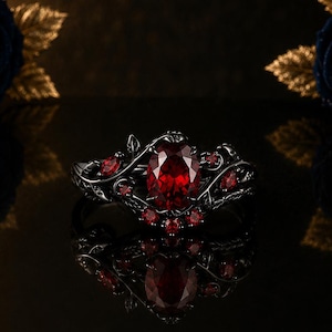 Puede incluir: Un anillo de metal negro con una piedra preciosa roja central de corte ovalado rodeada de piedras rojas más pequeñas. La banda presenta un intrincado diseño de metal en forma de enredadera que envuelve el dedo con gran detalle.