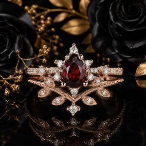 Op de afbeelding: Een roségouden ring met een peervormige, dieprode granaat als middensteen, omringd door kleine ronde witte accentstenen. De ringband heeft een bladvormig ontwerp met gedetailleerd metaalwerk en extra kleine witte steentjes in de zetting.