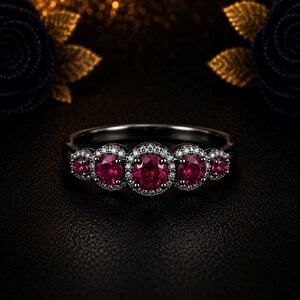 Puede incluir: Un anillo de metal negro con cinco piedras preciosas redondas de color rojo intenso dispuestas en fila. Cada piedra está rodeada por un halo de pequeñas piedras blancas transparentes. La piedra central es la más grande y disminuyen hacia los lados.