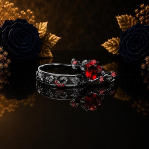 Könnte beinhalten: Ein Ring-Set aus schwarzem Metall, bestehend aus einem Band mit eingravierten Blattmustern und kleinen roten Steinen sowie einem passenden Ring mit einem herzförmigen roten Edelstein, umgeben von filigranen, rankenartigen Verzierungen.