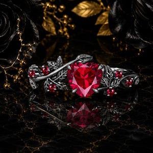 Pode incluir: Um anel de metal escuro com uma grande pedra preciosa vermelha em forma de coração no centro, ladeada por pedras vermelhas redondas menores incrustadas num motivo detalhado de videiras e folhas que envolve a banda do anel.
