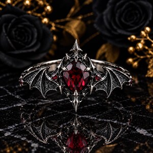 Puede incluir: Un anillo de tono plateado con una piedra preciosa central de color rojo intenso en forma de pera. La banda presenta motivos intrincados de alas de murciélago que se extienden desde el centro, con pequeñas piedras rojas redondas incrustadas.