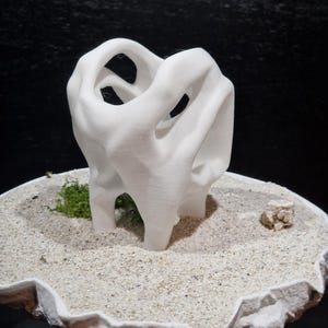 May include: A white, three-dimensional, abstract sculpture with a hollow, organic, and fluid form. The sculpture is positioned upright in a shallow, circular container filled with light-colored sand, a small patch of green moss, and a small rock.