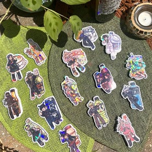 Puede incluir: Una colección de pegatinas holográficas con varios personajes de estilo anime combinados con diferentes recipientes de bebidas, incluyendo cartones de leche, botellas de plástico y latas, dispuestos sobre una superficie de tela verde texturizada.
