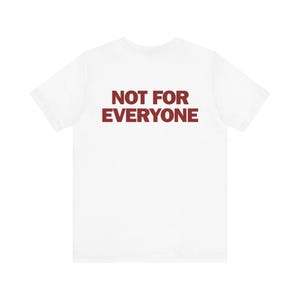 Può includere: Una t-shirt bianca a maniche corte con la scritta NOT FOR EVERYONE stampata in un carattere sans-serif rosso scuro in grassetto al centro del petto. Il capo presenta un girocollo standard e una struttura in tessuto bianco semplice.