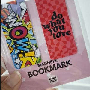 Puede incluir: Dos marcapáginas magnéticos en un paquete transparente. Uno presenta un diseño colorido estilo cómic con la palabra WOW. El otro es rojo con un patrón de cuadros y el texto negro do what you love. El paquete está etiquetado como MAGNETIC BOOKMARK.