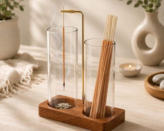 May include: An incense burner set featuring a rectangular wooden base with two clear glass cylinders. One cylinder holds a burning incense stick suspended from a gold-toned metal arm, while the other cylinder contains a bundle of unlit incense sticks.