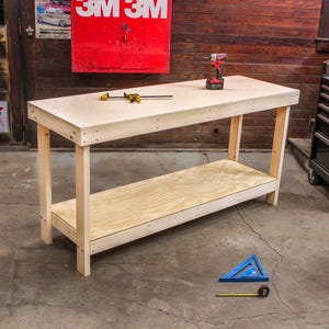 May include: A rectangular wooden workbench featuring a light-colored plywood top and a lower storage shelf supported by four sturdy legs. A red and black cordless drill and a yellow and black bar clamp rest on the surface of the workbench.