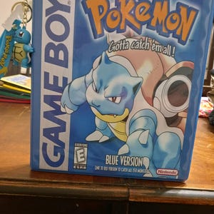 May include: A blue Nintendo Game Boy Pokémon Blue Version game case featuring Blastoise on the cover with the text Gotta catch 'em all! and an ESRB E rating. A small blue Squirtle character keychain is attached to the side of the game case.