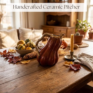 May include: A glossy, dark orange ceramic pitcher shaped like a gourd with a curved, textured handle. The pitcher features the text Handcrafted Ceramic Pitcher at the top. It is positioned on a wooden surface alongside a bowl of small gourds.