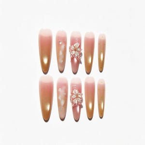 Könnte beinhalten: Ein Set aus zehn mandelförmigen Press-on-Nägeln mit einem Farbverlauf von Rosa zu Gold. Das Design umfasst zwei Nägel mit dreidimensionalen Blumenakzenten, zwei Nägel mit weißen blütenblattartigen Einschlüssen und zwei Nägel mit kleinen Goldpunkten.
