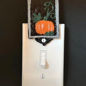 Op de afbeelding: Een witte lichtschakelaarplaat met een rechthoekige decoratieve opzet. De opzet heeft een zilveren lijst, een zwarte achtergrond en een verhoogde oranje pompoen met groene bladeren en ranken. Een zwarte sierbeugel verbindt de opzet met de plaat.
