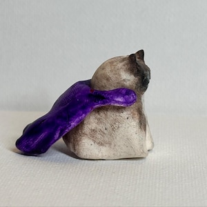 May include: A small ceramic figurine of a beige cat with dark brown markings on its face and ears. The cat is wearing a vibrant purple cape that drapes over its back and extends to the side, featuring a smooth, glossy finish on the fabric.
