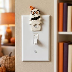 Op de afbeelding: Een witte rechthoekige lichtschakelaarplaat met een gebeeldhouwde spookfiguur aan de bovenkant. Het spook draagt een oranje heksenhoed, een zwarte bril en houdt een kleine zwarte vleermuis vast. De plaat heeft één tuimelschakelaar.