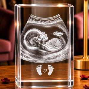 May include: A rectangular clear glass block featuring a laser-etched 3D ultrasound image of a fetus. Below the ultrasound, the glass displays two small baby footprints flanking a central heart icon, all etched in white.