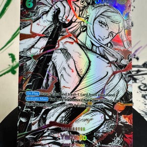 Könnte beinhalten: Eine holografische One Piece Sammelkarte mit der Figur Yamato. Die Karte zeigt eine schwarz-weiße Illustration im Manga-Stil mit roten Akzenten, Kosten von 6, 5000 Stärke und Text, der die Spielfähigkeiten wie On Play und Activate:Main beschreibt.