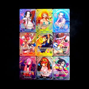 Puede incluir: Una colección de nueve cartas coleccionables de One Piece dispuestas en una cuadrícula de tres por tres. Cada carta presenta una colorida ilustración de un personaje de anime con texto impreso, incluyendo los nombres Boa Hancock, Nami, Vivi, Uta, Nico Robin, Perona, Reiju, Koala y Tashigi.