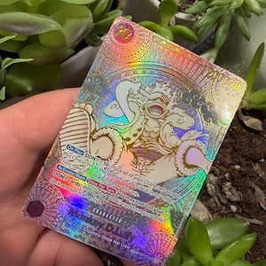 Puede incluir: Una carta coleccionable holográfica de One Piece que muestra a Monkey D. Luffy en forma Gear 5. La carta presenta intrincados patrones geométricos dorados, un nivel de poder de 12000 e instrucciones de juego detalladas en el texto.