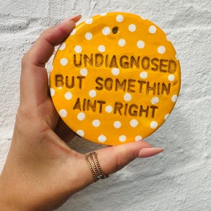 May include: A circular yellow ceramic plaque featuring white polka dots and the embossed text "UNDIAGNOSED BUT SOMETHIN' AINT RIGHT" in dark brown lettering. The plaque includes a small hole at the top for hanging purposes and has a glossy finish.
