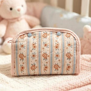 May include: A quilted pouch featuring a vertical striped pattern in light blue and cream, accented with small orange floral motifs. The pouch has a rounded top edge, a pink fabric trim, and two small blue circular buttons near the top closure.