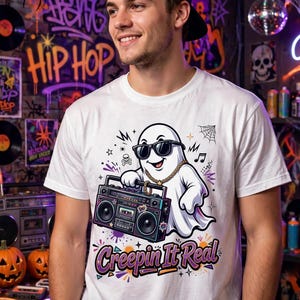 Puede incluir: Una camiseta blanca de manga corta con un gráfico de un fantasma de dibujos animados con gafas de sol negras y una cadena dorada, sosteniendo un radiocasete. El texto "Creepin It Real" aparece debajo en letras moradas y negras con detalles naranjas.