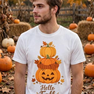 Può includere: Una t-shirt bianca a maniche corte con una grafica di tre zucche arancioni impilate, con la zucca centrale che indossa una sciarpa di lana marrone. La zucca in basso ha un volto intagliato. Il testo "Hello Fall" è stampato sotto in marrone.