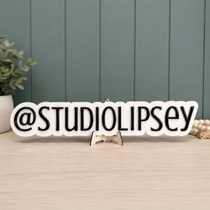May include: A white, custom-cut sign featuring the black text @STUDIOLIPSEY in a bold, sans-serif font. The sign is mounted on a small, light-colored wooden easel stand, displaying the handle clearly against a clean, contrasting background.