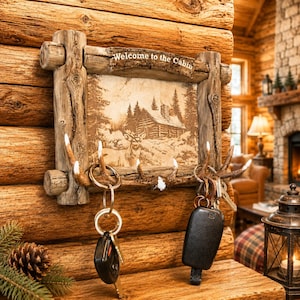May include: A rectangular wall-mounted key holder with a faux log frame and a central panel featuring a sepia-toned illustration of a cabin and a deer. The text Welcome to the Cabin is printed on the top frame. Antler-shaped hooks hold two key rings.