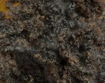 Op de afbeelding: Een close-up van een stapel bruine, zwarte en grijze wolvezels. De wol is zacht en pluizig en lijkt handgesponnen te zijn.