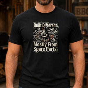 Könnte beinhalten: Ein schwarzes T-Shirt mit kurzen Ärmeln und einem Grafikdruck eines Mechanikers, der von mechanischen Zahnrädern und Werkzeugen umgeben ist. Der Text "Built Different." steht über der Illustration, "Mostly From Spare Parts." steht darunter.