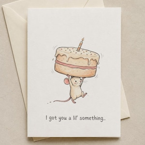 May include: A white greeting card featuring a cartoon illustration of a small beige mouse lifting a large cake with pink filling and a single lit candle on top. The text below the mouse reads, I got you a lil' something..