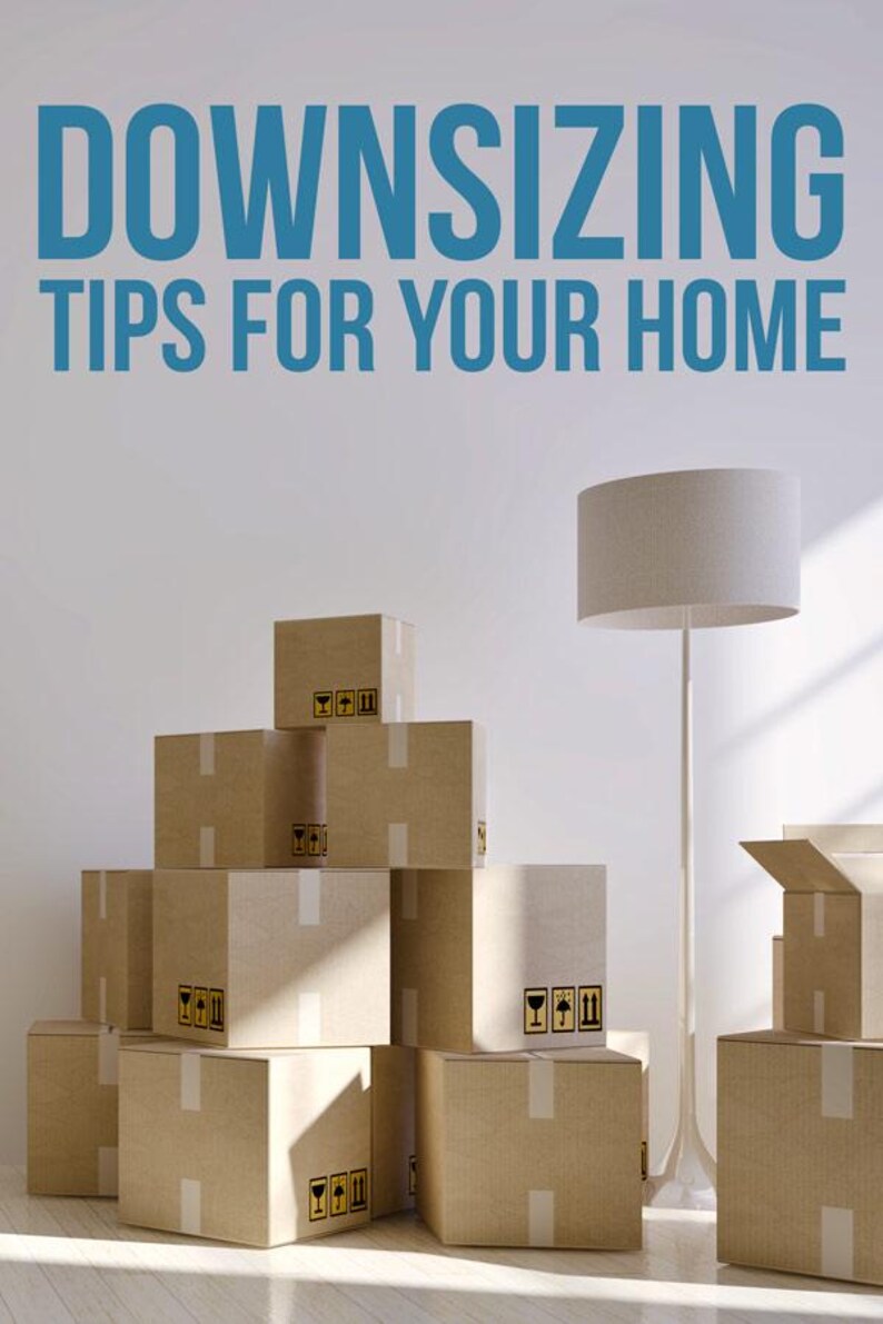 May include: A stack of brown cardboard moving boxes with white tape and black fragile symbols, positioned next to a tall white floor lamp. The text DOWNSIZING TIPS FOR YOUR HOME is printed in large blue letters at the top of the image.