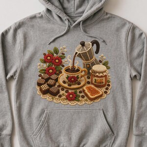 Puede incluir: Una sudadera con capucha gris jaspeado que presenta un gráfico central de una escena de café tejida a ganchillo. El diseño incluye una cafetera moka vertiendo café en una taza, un tarro de mermelada, tostadas y chocolates sobre un tapete beige.