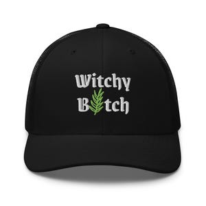 Può includere: Un cappellino trucker nero con testo ricamato in bianco che recita Witchy Bitch, con un rametto di rosmarino verde ricamato che sostituisce la lettera i nella seconda parola. Il cappellino ha un pannello frontale strutturato e rete.