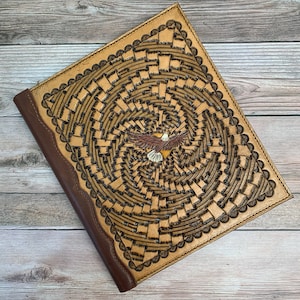 May include: A brown leather journal with a detailed eagle design and a woven pattern. The journal has a brown leather spine and a decorative edge.