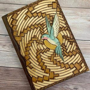 May include: A brown leather journal with a woven pattern and a hummingbird design. The hummingbird is painted in shades of blue, green, and pink.