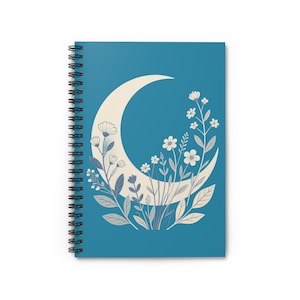 Puede incluir: Un cuaderno de espiral con una cubierta azul profundo que presenta un gráfico grande de una luna creciente de color crema. La luna está decorada con un grupo de flores silvestres y hojas ilustradas de colores claros que crecen hacia arriba.