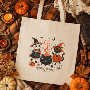 Puede incluir: Una bolsa de tela beige con un gráfico de dos gatos vestidos de brujos junto a un caldero negro con vapor naranja. El diseño incluye estrellas, una luna creciente, pequeñas calabazas y el texto Spooky Season en letra cursiva.