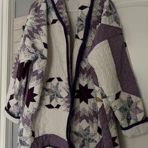 Puede incluir: Una chaqueta acolchada con un patrón de estrellas en tonos morados, blancos y estampados florales. La prenda tiene un cuello tipo chal, mangas largas con puños doblados y un ribete morado oscuro en los bordes, confeccionada con costuras visibles.