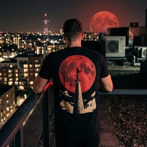 May include: A black t-shirt featuring a graphic print on the back. The design displays a large red moon behind a tall, pointed sword standing vertically. A small silhouette of a figure stands atop the sword, with stylized clouds at the base.