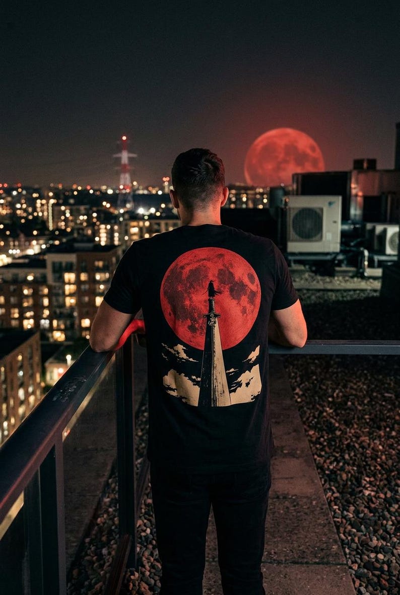 May include: A black t-shirt featuring a graphic print on the back. The design displays a large red moon behind a tall, pointed sword standing vertically. A small silhouette of a figure stands atop the sword, with stylized clouds at the base.