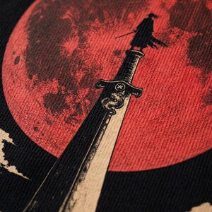 May include: A black t-shirt featuring a graphic print of a large red moon in the background. In the foreground, a silhouette of a person stands atop a long sword pointing upward. The sword blade is decorated with a snake emblem and a small symbol.