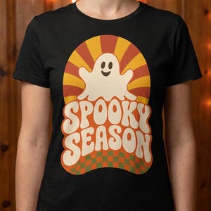 May include: A black short-sleeve t-shirt featuring a graphic print of a smiling white ghost centered over a yellow and orange sunburst pattern. Below the ghost, the text SPOOKY SEASON is printed in large white letters over a green and orange checkerboard.