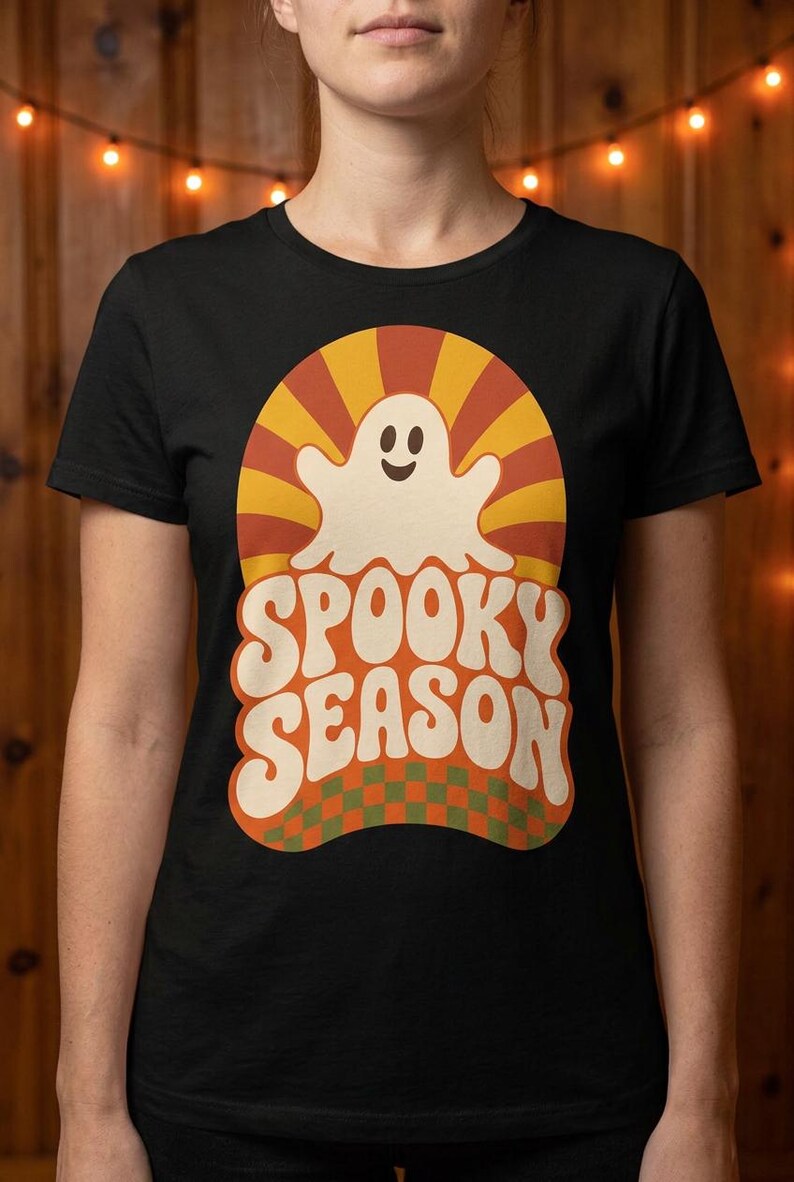 May include: A black short-sleeve t-shirt featuring a graphic print of a smiling white ghost centered over a yellow and orange sunburst pattern. Below the ghost, the text SPOOKY SEASON is printed in large white letters over a green and orange checkerboard.